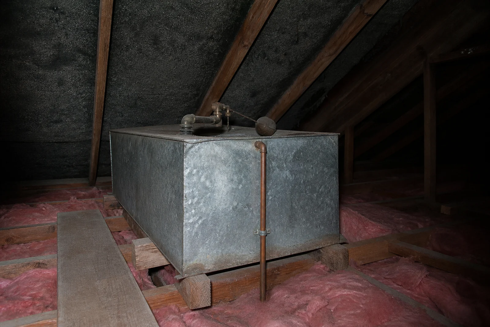 A galvanised header tank sitting on timber framing in a roof space with a pipe running down from it. A header tank in the roof is the clearest sign that a house is on a low pressure hot water system.