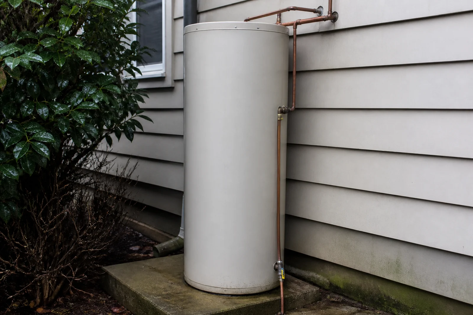 A hot water cylinder installed outdoors against the weatherboard wall of a New Zealand house, standing on a level concrete base with its pipework and relief valve drain visible.